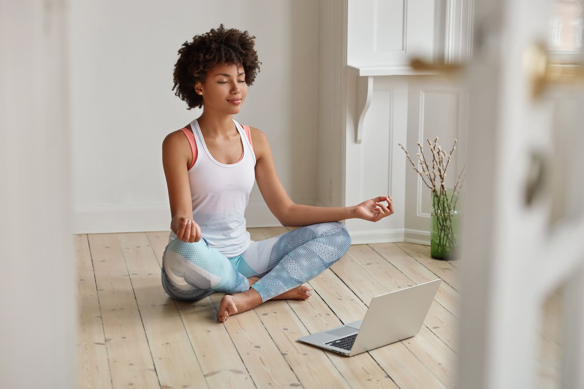 relaxed-dark-skinned-woman-with-sporty-body-sits-in-zen-pose-keeps-legs-crossed-watches-yoga-lessons-on-laptop-computer-using-internet-meditates-on-floor-in-empty-room-wellness-concept